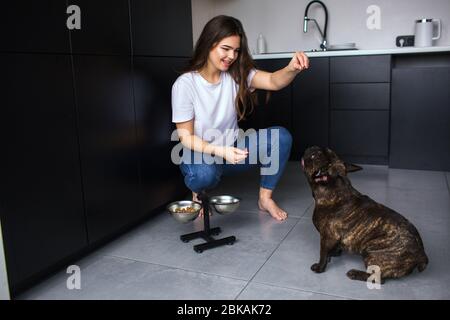 Giovane donna in cucina durante la quarantena. Ragazza sedersi e giocare con il bulldog francese. Animale domestico di addestramento per fare i comandi e mangiare l'alimento del cane dopo quello. Foto Stock