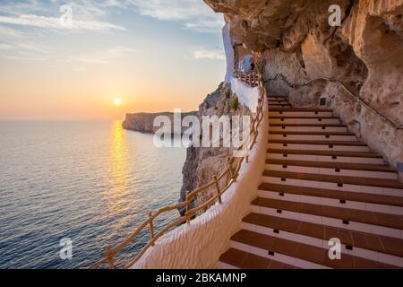 Tramonto costiero su una scala a Cova d'en Xoroi, un bar e night club situato in grotte naturali, Cala en Porter, Minorca, Spagna Foto Stock