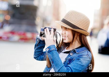 Ragazza che indossa un cappotto in denim, una sciarpa arancione e jeans che cammina per il centro della città, scattando foto e porta un orso giocattolo sulle spalle Foto Stock