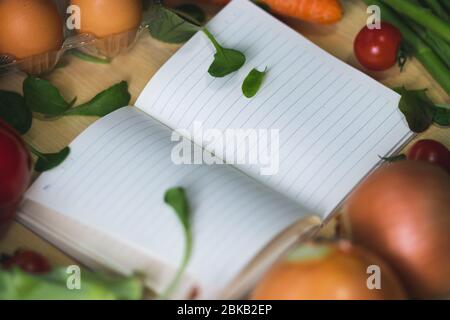 Notebook aperto circondato da verdure. Lista della drogheria con verdure intorno esso. Lista di acquisti con verdure che la circondano. Shopping per i generi alimentari Foto Stock