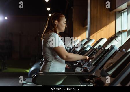 vista laterale della messa a fuoco della ragazza sovrappeso sulla pedana mobile in palestra Foto Stock