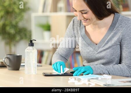 Donna felice che pulisce lo smartphone con igienizzatore e tessuto che impedisce il covid-19 con guanti in lattice seduti su una scrivania a casa Foto Stock