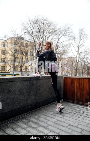 Bella ragazza bionda con coda di ponytail è l'allenamento in abiti sportivi per le strade di una città. Indossa una giacca sportiva, leggings e sneaker. Foto Stock