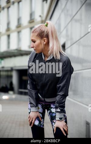 Bella ragazza bionda con coda di ponytail è l'allenamento in abiti sportivi per le strade di una città. Indossa una giacca sportiva, leggings e sneaker. Foto Stock
