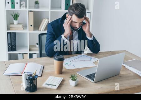 uomo d'affari stressato e sfinito che tocca la testa mentre si siede con gli occhi chiusi sul posto di lavoro vicino a documenti e laptop Foto Stock