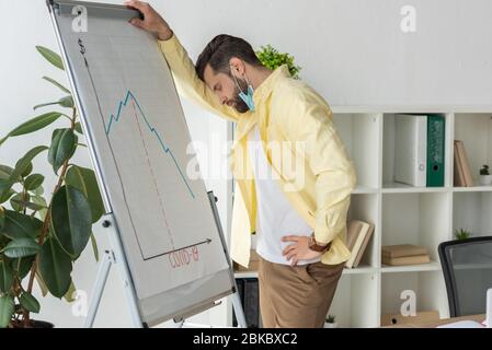 uomo d'affari stressato in piedi con gli occhi chiusi vicino alla lavagna a fogli mobili con l'iscrizione covid-19 e grafici che mostrano diminuzione Foto Stock