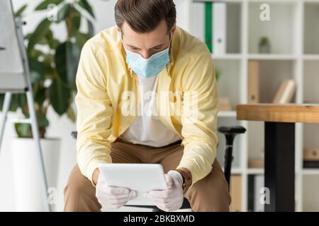 uomo d'affari in maschera medica e guanti in lattice con tablet digitale in ufficio Foto Stock