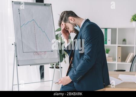 vista laterale dell'uomo d'affari stressato in piedi con la testa piegata e gli occhi chiusi vicino alla lavagna a fogli mobili con grafici che mostrano la recessione Foto Stock