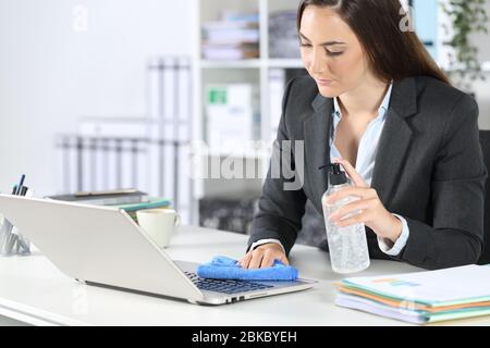 Donna executive che disinfetta il laptop con igienizzatore e panno seduto su una scrivania in ufficio Foto Stock