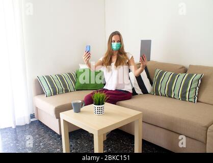 giovane donna in una maschera medica autoisolata a casa è seduta su un divano con uno smartphone e un tablet in entrambe le mani Foto Stock