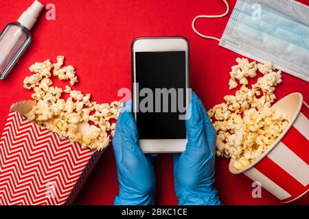 Vista dall'alto della persona che tiene lo smartphone con schermo vuoto vicino alla maschera medica e popcorn sulla superficie rossa Foto Stock