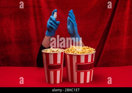 Vista tagliata della donna in guanti di lattice che tiene bottiglia di igienizzante per le mani vicino popcorn su superficie rossa con velluto sullo sfondo Foto Stock