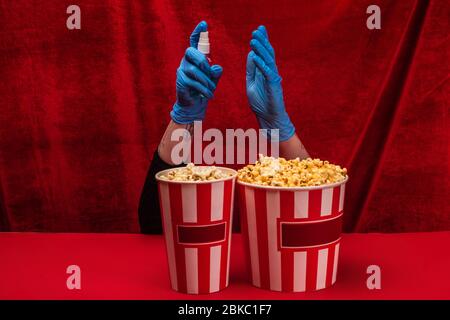 Vista tagliata del popcorn vicino alla donna in guanti di lattice che tiene igienizzante mani su superficie rossa con velluto sullo sfondo Foto Stock