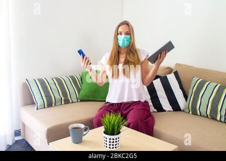 una giovane donna in una maschera medica in quarantena è seduta con occhi sorpresi con un telefono e tablet in entrambe le mani Foto Stock