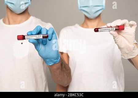 Vista tagliata della coppia in maschere mediche e guanti in lattice contenenti provette con campioni di sangue e scritte di coronavirus isolate in grigio Foto Stock