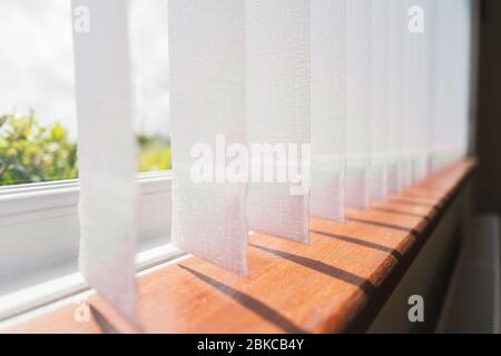 Tende per finestre verticali bianche con tasche senza fili incollate e pesate sull'estremità che gettano ombre sul davanzale della finestra in legno. Foto Stock