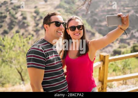 Coppia felice di viaggio facendo selfie montagne sfondo, colori estivi soleggiati, umore romantico. Occhiali da sole eleganti. Felici risate facce emotive. Foto Stock