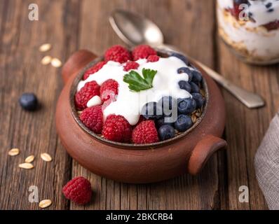 Granola con yogurt bianco con lamponi e mirtilli in ciotola in ceramica su scrivania in legno naturale. Foto Stock