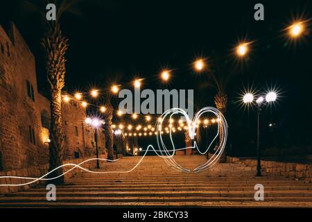 Una luce a forma di cuore che si disegna su un lungo passo sui gradini di Jaffa. Prospettiva di lanterne e ghirlande con bulbi. Sullo sfondo di Palm tre Foto Stock