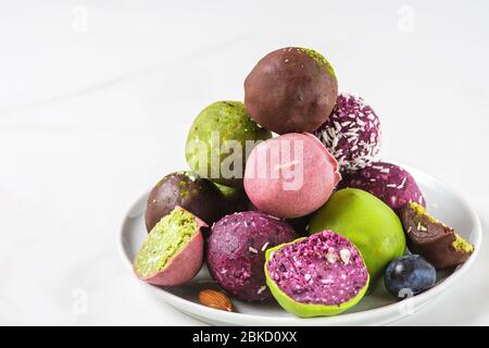 Cibo sano vegano da dessert. Palle energetiche colorate e crude in glassa di cioccolato in un piatto su tavolo bianco. Spuntini deliziosi Foto Stock