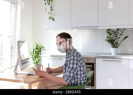 Uomo freelancer in occhiali che indossano una maschera protettiva medica, guardando lo smartphone e scrive le note della penna in un notebook, lavorando online da casa durante l' Foto Stock