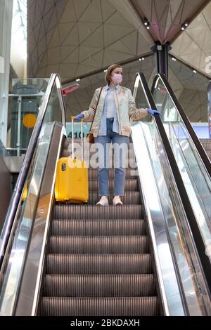 Donna con bagagli gialli si trova sulla scala mobile presso il terminal dell'aeroporto quasi vuoto a causa di restrizioni di viaggio per l'epidemia di coronavirus/Covid-19. Quar Foto Stock