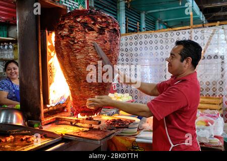 Cameriere che taglia porzioni di carne da un grande shawarma che sta cucinando sopra un fuoco in un ristorante di strada nel mercato comunale di Merida. Foto Stock