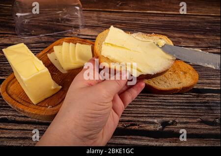 Qualcuno spalma un po' di burro con un coltello su pane tostato di grano fresco, un pezzo di burro in un piatto di burro di legno e un pane tostato di grano fresco su uno schienale di legno Foto Stock