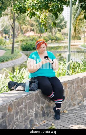 Giovane donna di dimensioni più poggiata su un muro di pietra nel parco e controllando messaggi e notifiche nel suo smartphone Foto Stock