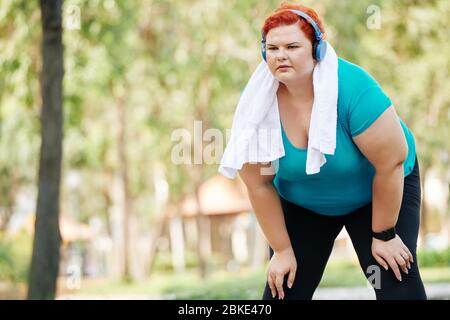 Donna stanca e di dimensioni più giovani con asciugamano su spalla riposante dopo jogging in parco Foto Stock