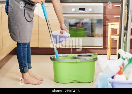 Casalinga in grembiule versando il liquido di pulizia del pavimento con profumo di lavanda in secchio con mop Foto Stock