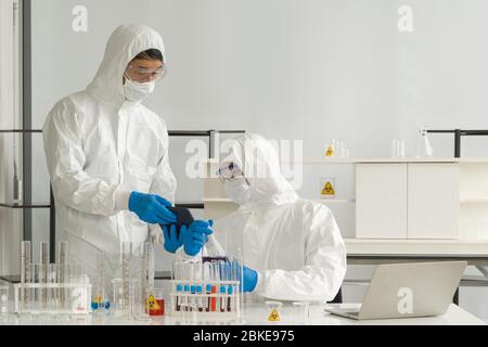 I ricercatori epidemiologici in abbigliamento protettivo virus utilizzano i computer tablet per la ricerca di composti chimici su Internet. Atmosfera di lavoro in ch Foto Stock