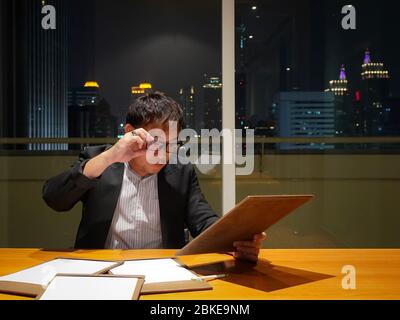 Un uomo d'affari asiatico esamina un breve documento dopo il lavoro. Sala riunioni ai piani alti di notte. La vivace area del centro di Bangkok. Foto Stock