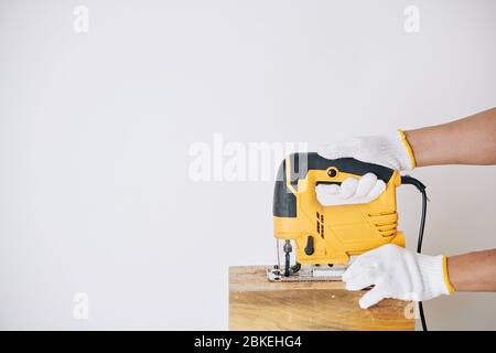 Immagine ravvicinata del falegname in guanti protettivi per tagliare la tavola di legno con un seghetto elettrico Foto Stock