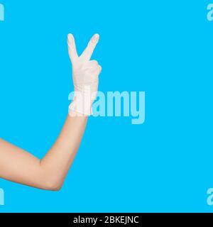 Gesto di pace della mano. Segno di vittoria. Indossare un guanto monouso bianco in lattice per uso medico isolato su blu. Gesto della mano della donna o segno isolato su bianco. Foto Stock