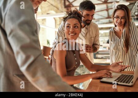 Lavorare insieme. Giovane e allegra donna afro americana che usa il laptop e discute qualcosa con i colleghi mentre si siede nell'ufficio moderno Foto Stock