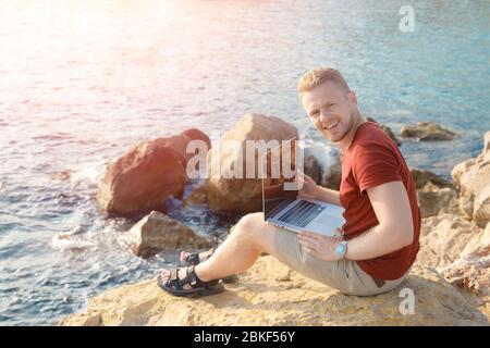Ragazzo sorridente in pantaloncini che si siedono su una grande roccia sulla costa con un notebook. Freelancing, concept tele work, business di successo Foto Stock