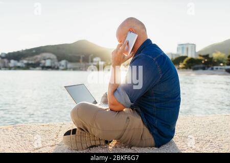 uomo che parla al telefono e che usa il laptop sulla spiaggia al tramonto. Uomo che lavora in remoto online utilizzando un notebook seduto in spiaggia all'ora del tramonto. Foto Stock