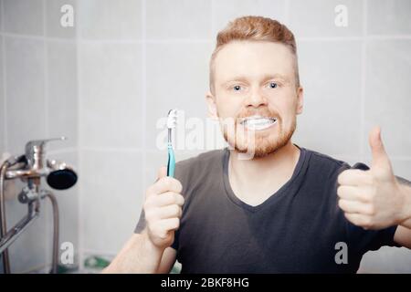 L'uomo con la barba spazzola i denti in dentifricio e spazzola in bagno. Mostra il pollice perfettamente. Foto Stock