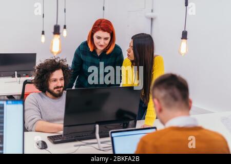 Due giovani donne d'affari accanto a un collega maschile mentre spiega le nuove attività online su un computer in un ambiente di ufficio confortevole. Foto Stock