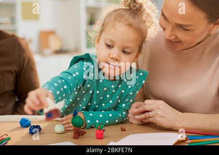 Ritratto medio orizzontale di bambina che indossa il vestito turchese di pois polka seduta al tavolo con i genitori che fanno le forme usando l'impasto colorato del gioco Foto Stock