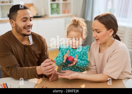 Giovani genitori seduti a tavola aiutando la loro figlia piccola a fare colorful modeling palla di argilla, colpo orizzontale Foto Stock