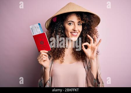 Bella donna turistica con capelli ricci e piercing indossare cappello asiatico tenendo passaporto giappone facendo ok segno con le dita, simbolo eccellente Foto Stock