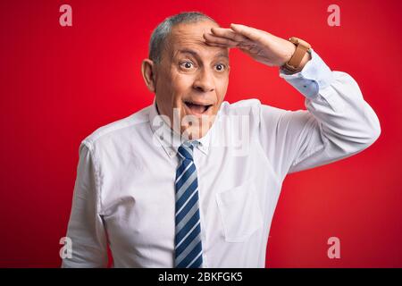 Uomo d'affari anziano che indossa la cravatta elegante che sta levandosi in piedi sopra il fondo rosso isolato molto felice e sorridente guardando lontano via con mano sopra la testa. Mare Foto Stock