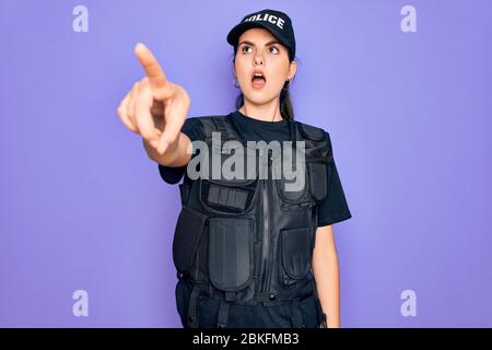 Giovane donna di polizia indossato uniforme giubbotto di sicurezza a prova di proiettile su sfondo viola puntando con il dito sorpreso davanti, bocca aperta espressione stupita Foto Stock