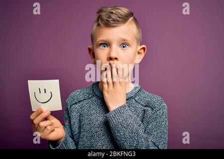 Giovane capretto caucasico che mostra faccia smiley su una nota di carta come felice messaggio coprire bocca con mano scioccata con vergogna per errore, espressione di fe Foto Stock