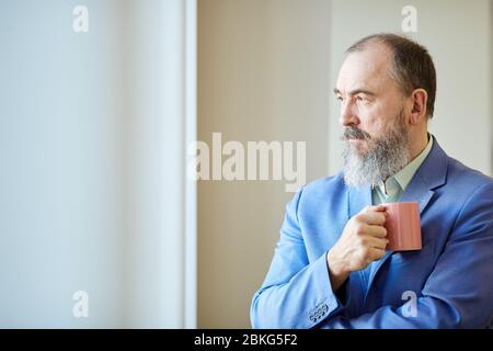 Uomo anziano elegante con barba sul viso e abito blu che tiene una tazza di caffè guardando fuori dalla finestra, spazio per le copie Foto Stock