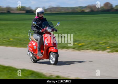 Kiel, Germania. 04 maggio 2020. In tempo soleggiato, una donna guida il suo scooter rosso Vespa 125 cc lungo una strada sterrata indossando indumenti di sicurezza, casco e guanti. Dal 01.01.2020, i titolari di una patente di guida non hanno più bisogno di una patente di guida per motocicli separata. Sono sufficienti solo 5 ore di guida e 4 ore teoriche per poter guidare veicoli a due ruote fino a 125 ccm. Credit: Axel Heimken/dpa/Alamy Live News Foto Stock