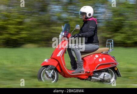 Kiel, Germania. 04 maggio 2020. In tempo soleggiato, una donna guida il suo scooter rosso Vespa 125 cc lungo una strada sterrata indossando indumenti di sicurezza, casco e guanti. Dal 01.01.2020, i titolari di una patente di guida non hanno più bisogno di una patente di guida per motocicli separata. Sono sufficienti solo 5 ore di guida e 4 ore teoriche per poter guidare veicoli a due ruote fino a 125 ccm. Credit: Axel Heimken/dpa/Alamy Live News Foto Stock