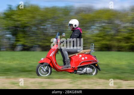 Kiel, Germania. 04 maggio 2020. In tempo soleggiato, una donna guida il suo scooter rosso Vespa 125 cc lungo una strada sterrata indossando indumenti di sicurezza, casco e guanti. Dal 01.01.2020, i titolari di una patente di guida non hanno più bisogno di una patente di guida per motocicli separata. Sono sufficienti solo 5 ore di guida e 4 ore teoriche per poter guidare veicoli a due ruote fino a 125 ccm. Credit: Axel Heimken/dpa/Alamy Live News Foto Stock